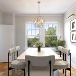 Bright dining room with a rectangular table, six upholstered chairs, a modern pendant light, framed abstract art on the wall, and two large windows with a view of green trees.