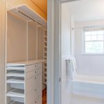 A bright walk-in closet with white drawers and shelves adjoins a bathroom featuring a bathtub, white tile floor, towel rack, and a window letting in natural light.