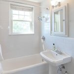 A bright, clean bathroom with white tile walls, a bathtub with a white towel, a pedestal sink, a wall mirror, and a window letting in natural light.