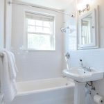 Bright, clean white bathroom with a bathtub, pedestal sink, wall mirror, towel rack with white towels, wall-mounted light, toilet paper holder, and a window letting in natural light.