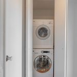 A stacked washer and dryer set is tucked inside a small closet with white double doors, partially open. The floor is hardwood with a parquet pattern, and the closet is lit by a ceiling light.