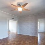 A bright, empty room with parquet wood flooring, a ceiling fan with a light, white walls with one textured accent wall, large windows, and two doorways leading to other rooms.