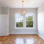Bright, empty room with light gray walls, parquet wood floor, a modern spherical ceiling light, two large windows with a view of trees, and a door leading to a tiled bathroom.