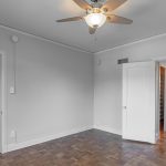 A bright, empty bedroom with parquet floors, light gray walls, a ceiling fan with a light, two white doors, and an adjoining bathroom visible through one open door.