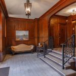 Elegant building lobby with wood-paneled walls, a cushioned cream sofa, a side table with a lamp, framed landscape painting, ornate lighting fixtures, and tiled stairs with a black wrought-iron railing.