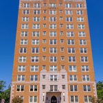 A tall, tan brick building with many windows stands under a clear blue sky. The entrance has an arched doorway and a sign reading "Forest Park Tower" above manicured landscaping and a sidewalk.