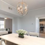 A modern dining room with a light wood table, beige chairs, and a vase of white tulips. A unique geometric chandelier hangs above. In the background are a living room with a sofa and a kitchen with built-in appliances.
