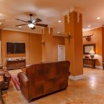 A spacious living room with tan walls and brown leather furniture, featuring a TV on the wall, a ceiling fan, rustic decor, a round dining table with chairs, and warm lighting on polished floors.