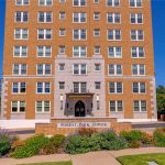 A multi-story brick apartment building with white trim, labeled "Forest Park Tower" on a sign in front, surrounded by landscaped bushes, flowers, and a clear blue sky.