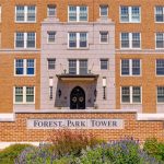 A brick apartment building with many windows, a central entrance, and a sign in front reading "Forest Park Tower." Colorful plants and flowers are landscaped around the sign.