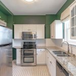 A kitchen with white cabinets, granite countertops, stainless steel appliances, a double oven, a refrigerator, green walls, white tile backsplash, and a window above a double sink. The floor has a patterned tile design.