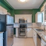 A kitchen with green walls, white cabinets, stainless steel appliances, granite countertops, and a patterned tile floor. A window is above the sink, and a round ceiling light is overhead.