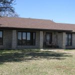 Single-story brick house with a brown shingle roof, large windows, and a covered front porch with a bench swing. The front yard has sparse grass, with trees and a clear blue sky in the background.