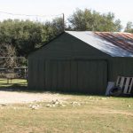 A green metal shed with a rusted roof stands beside a dirt road, with scattered equipment, a tire, and a pallet leaning against it. Trees and old machinery are visible in the background.