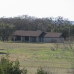 A single-story house with a dark roof sits on a large, grassy plot surrounded by trees; a white truck is parked on the dirt driveway nearby under a clear sky.