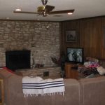 A cozy living room with a large stone fireplace, a brown sectional sofa, a ceiling fan, wood-paneled walls, and a TV displaying a black-and-white show. Blankets and clothes are piled on the sofa.