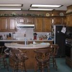A spacious kitchen with wooden cabinets, a central island with four wooden chairs, various kitchen appliances, and decorative plates on the upper shelves. The room is well-lit with fluorescent ceiling lights.