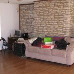 A living room with a stone accent wall, beige couch with boxes on it, a desk with an old computer, a green cabinet, wood floors, and a saddle on a stand in the corner. Light filters through closed blinds.