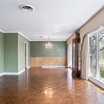 A spacious living room with parquet wood floors, green and beige walls, a large picture window with a view of trees outside, a chandelier, built-in bookshelf, and a glass-front corner cabinet.