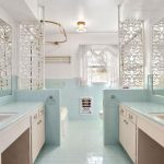 A vintage bathroom with pastel blue tiles, double sinks, large mirrors, lace curtains on windows, brass fixtures, and ornate white decorative screens on either side of the bathtub.