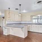 Spacious kitchen with vintage cream cabinets, granite countertops, pendant lights, and parquet flooring. Open layout leads to a living area with a brick fireplace and large windows.