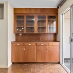 A built-in wooden cabinet with glass doors and lower drawers stands against a wall near sliding glass doors, next to a partially open, cream-colored partition wall in a room with parquet flooring.