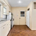 A kitchen with white cabinets, a double sink, granite countertops, dishwasher, oven, and refrigerator. The floor is patterned wood, and there are large windows above the sink and a door at the back.