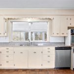 Vintage kitchen with cream-colored cabinets, a wide window above a double sink, granite countertops, a built-in double oven, and a dishwasher, all set on a wood parquet floor.