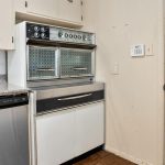 Vintage kitchen with a built-in double oven featuring retro dials and patterns, white cabinets, granite countertop, stainless steel dishwasher, wall thermostat, and a beige door with a round black handle.