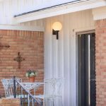 A small porch with a white metal table and chairs, a potted plant on the table, brick walls, a railing by the steps, and a glass door. A round light fixture is mounted on the white siding.