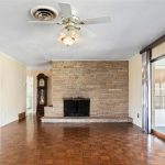 A spacious living room with parquet wood flooring, a stone fireplace, ceiling fan with light, and large sliding glass doors opening to an enclosed patio area. Adjacent rooms and a grandfather clock are also visible.