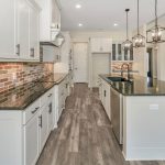 Modern kitchen with white cabinets, dark countertops, brick backsplash, stainless steel sink in a large island, pendant lights, and wood-look flooring in an open-concept layout.