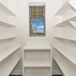 A white walk-in pantry with built-in shelves on both sides, a small shelving unit at the end, and a window with blinds letting in natural light.