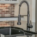 A modern kitchen sink with a black pull-down spring faucet on a shiny dark countertop. In the background, there are white cabinets and a brick-patterned backsplash.