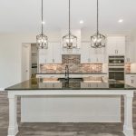 Modern kitchen with a large island, black countertop, white cabinets, brick backsplash, stainless steel appliances, three pendant lights, and gray wood-look flooring. The space is bright with ample natural light.