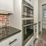 A modern galley kitchen with white cabinets, a brick backsplash, dark countertops, stainless steel wall ovens, and wood-look flooring. A small pantry with shelving is visible through an open door at the end.