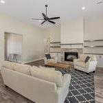 Spacious living room with light walls, gray wood flooring, cream sofas, patterned rug, brick fireplace, built-in shelves, ceiling fan, and large windows with blinds letting in natural light.