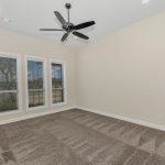 A bright, empty room with beige walls, three large windows with white blinds, brown carpet, a ceiling fan with black blades, and a sliding barn door partially open on the left.