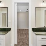 Modern bathroom with two white vanities, black countertops, rectangular mirrors, and black fixtures. A doorway in the center leads to a carpeted room with a ceiling fan. Bright lighting above each mirror.