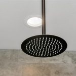 Close-up of a round, black rainfall showerhead mounted on the ceiling, with gray tiled walls and a recessed ceiling light in the background.