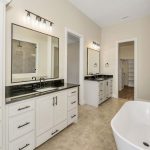 Modern bathroom with double vanities, black fixtures, white cabinets, a large freestanding bathtub, beige tile floor, and walk-in closet visible through an open doorway.