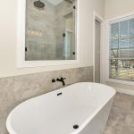 Modern bathroom with a white freestanding bathtub, wall-mounted black faucet, large mirror, a glass shower, tiled floors and walls, and a window with white blinds letting in natural light.