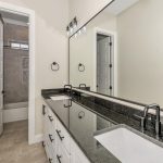 Modern bathroom with a long black countertop, double sinks, black fixtures, and white cabinets. A large mirror runs above the vanity. In the background, there is a bathtub and a doorway with white paneled doors.