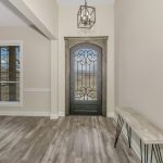 Entryway with a black ornate glass front door, light wood flooring, a rustic bench against the wall, and a dining area with a wooden table and white chairs to the left. A window with blinds is also visible.