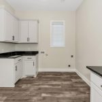 Modern laundry room with white cabinets, black countertops, wood-look flooring, beige walls, and a window with closed blinds. There are hookups for a washer and dryer along one wall.