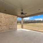 Covered patio with a ceiling fan, brick walls, and concrete floor overlooking a grassy yard with trees in the distance under a partly cloudy blue sky.