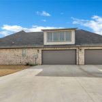 A modern brick house with a dark gray roof features three large garage doors and a spacious driveway under a bright blue sky with scattered clouds. The lawn is neatly trimmed and there are no cars parked outside.