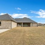 A single-story brick house with a large shingled roof, spacious backyard, and concrete patio area under a blue sky with scattered clouds. Leafless trees and another house are visible in the distance.