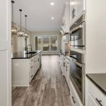 Modern kitchen with light cabinets, double wall ovens, black countertops, island with pendant lights, wood-style flooring, and large windows in the background letting in natural light.
