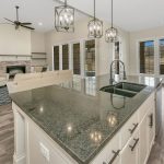 Modern open-concept kitchen and living room with a large island, pendant lights, stainless steel sink, beige sofas, fireplace, built-in shelves, and many windows allowing natural light.